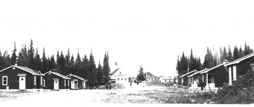 Les premières petites maisons situées en bas de la côte, près du lac. ON y voit aussi l'église et le premier presbytère. On y voit des arbres et des personnes marcher sur le chemin où sont situées de part et d'autre les maisons et qui mène au lac Simon.