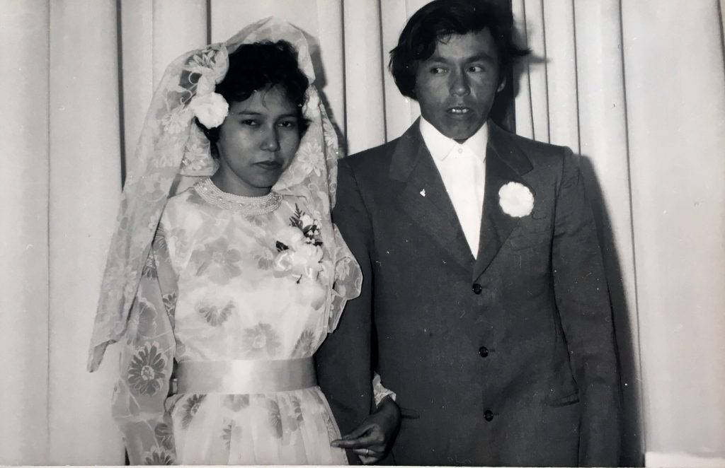 Un couple de jeunes mariés anicinabek le jour de leur mariage. L'homme porte une complet. La femme porte une robe blanche à fleurs en plus d'un voile dans ses cheveux et d'une bouquetière. Photo en noir et blanc. 