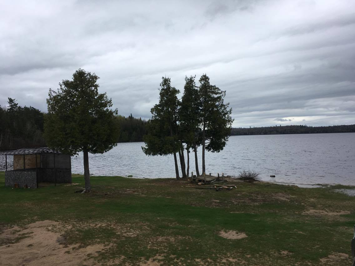 Site culturel de la communauté situé au bord de l'eau. On y voit quelques arbres, une petite portion de plage, le lac et de la forêt au loin. Le ciel est sombre. Une petite cabane où l'on fait fumer de la viande est sur la droite. Photo en couleur.