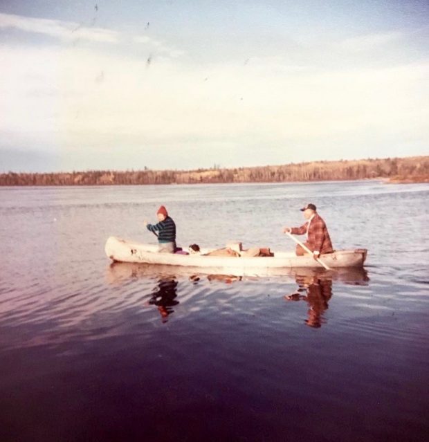 Un couple se déplace en canot sur un lac. Les deux rames à l'aide de pagaies. L'homme est à l'arrière, la femme à l'avant. La forêt est au loin. Photo en couleur. 