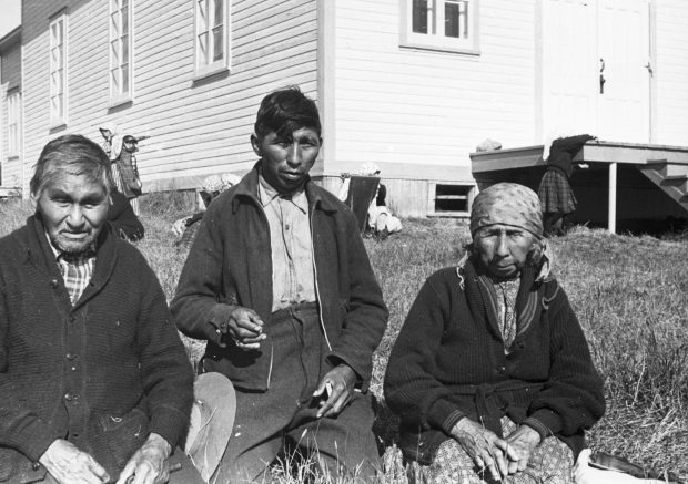 Une famille anicinabe est assise dans l'herbe près du Lac Simon. De gauche à droite: un homme âgé, un homme plus jeune (le fils) et une femme âgée. Ils portent des vêtements longs (vestes, chemises, pantalons, robe) et la femme a un foulard sur ses cheveux. L'église est derrière eux. On y voit aussi des enfants et une femme. Photo en noir et blanc.