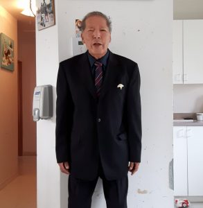 Portrait d'un ancien chef de Lac Simon. Habillé d'un complet avec cravate, il se tient debout dans sa cuisine. Photo en couleur. 