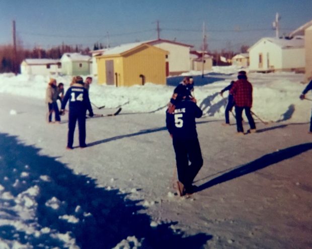 Des jeunes anicinabek jouant au hockey dans les rues de la communauté. La route est enneigée et de la neige borde les allées. On y voit quelques maisons aux alentours. Photo en couleur.
