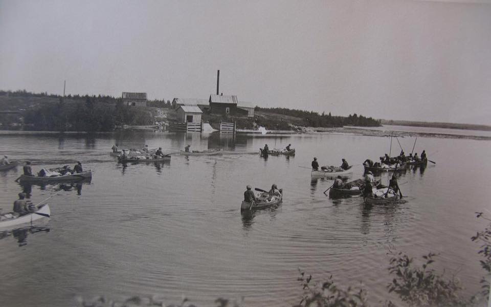 Arrivée des familles en canot au rassemblement d'été. On y voit une douzaine de canots avec des personnes à bord et des bagages. Sur la rive, on voit quelques petites maisons. Photo en noir et blanc.