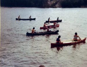 Canoe race at Lac Simon. We see five canoes with two people on board each of them. They were paddling. Some are wearing life jackets, some are not. Picture in color.