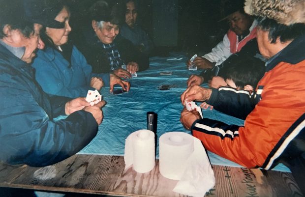 Groupe d'aînés anicinabek jouant aux cartes. Sept d'entre eux sont attablés avec des cartes en mains. Ils portent des manteaux et des chapeaux. Photo en couleur. 