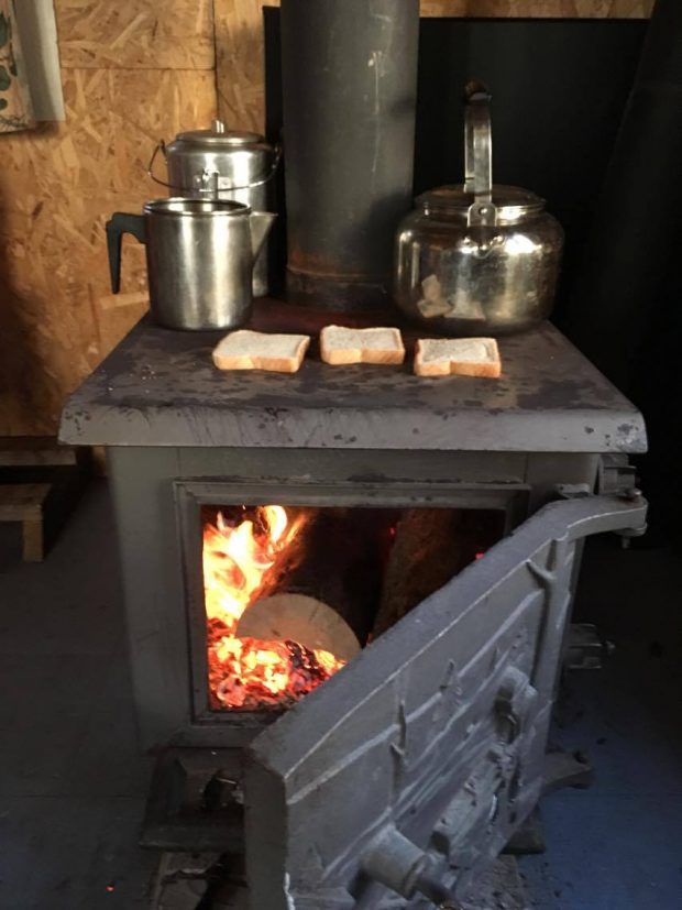 Du thé et des rôties chauffent sur un poêle a bois dont la porte est ouverte, ce qui laisse voir le feu. Trois théières et trois tranches de pains sont à cuire sur le dessus du poêle. Photo en couleur.