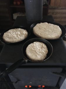 Trois banniques cuisent dans des poêlons posés directement sur un pôle à bois. Photo en couleur.