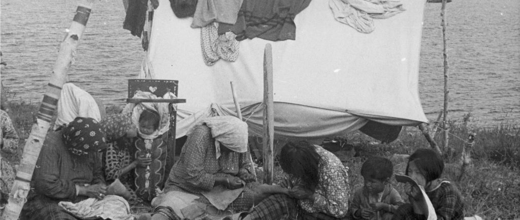 Des femmes anicinabek sont assises au bord du Lac Simon. Elles sont en train de coudre. Des enfants les regardent. Une tente, sur laquelle sont placés des tissus, est en arrière plan La photo date du milieu du 20e siècle. Photo en noir et blanc.