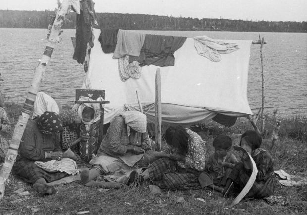 Des femmes anicinabek sont assises au bord du Lac Simon. Elles sont en train de coudre. Des enfants les regardent. Une tente, sur laquelle sont placés des tissus, est en arrière plan La photo date du milieu du 20e siècle. Photo en noir et blanc.