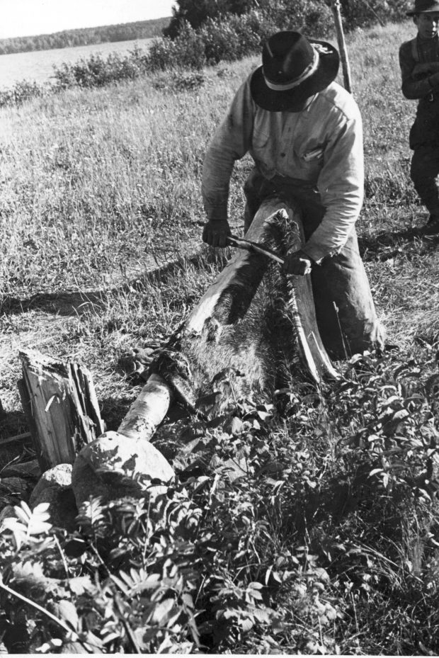 Un homme, qui porte un chapeau et des vêtements longs, travaille une peau d'animal avec un grattoir. La peau est installée sur un support de bois. Un autre homme est derrière lui. Ils sont sur le bord d'un lac. Photo en noir et blanc.