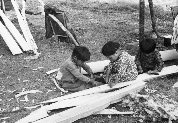 Trois jeunes anicinabek qui participent à la confection d'un canot en étant assis au sol. Ils utilisent des outils sur des retailles de bois. Photo en noir et blanc.