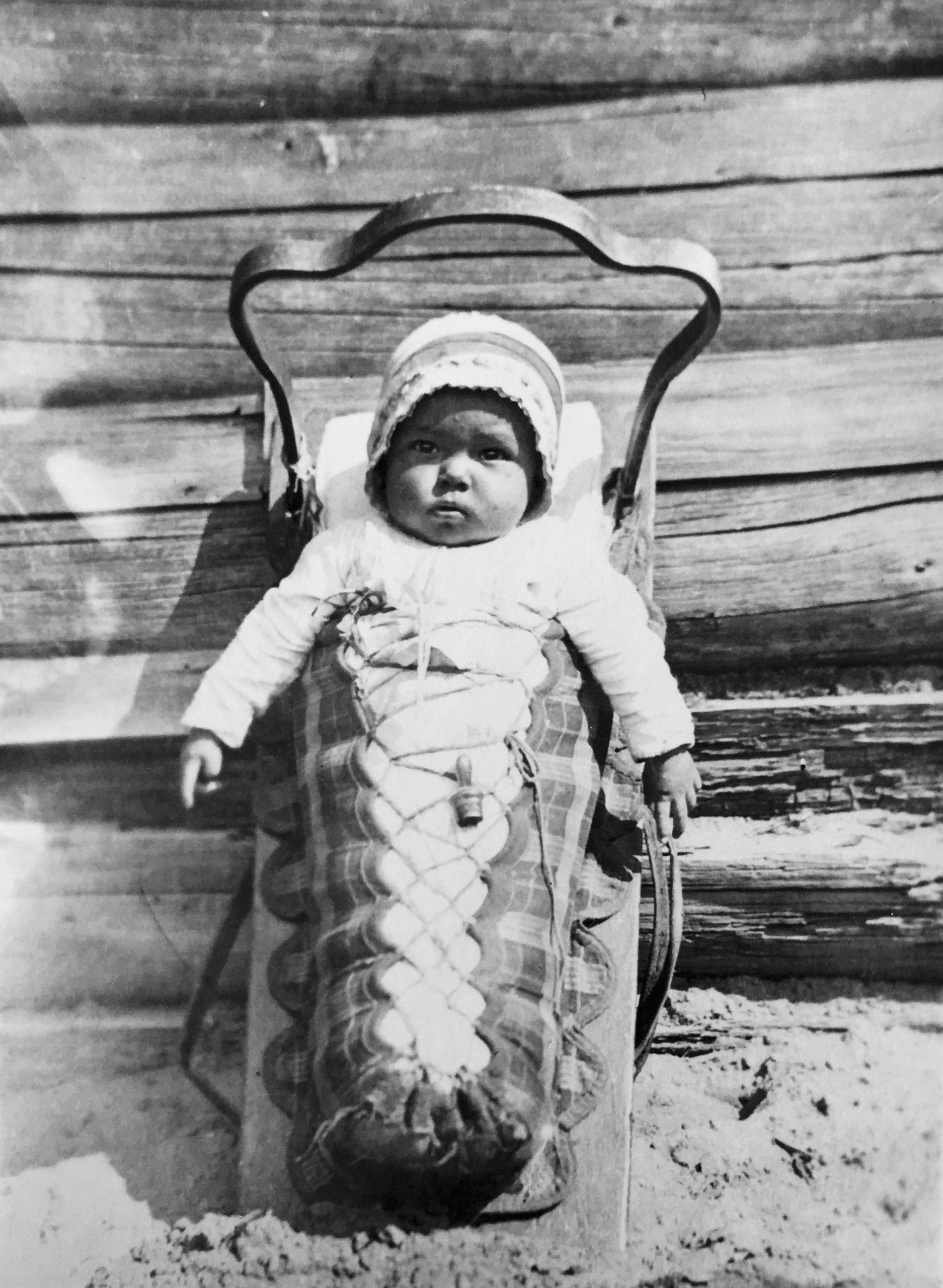 Un bébé dans son porte bébé (tikinagan). Il porte un chapeau. Ses bras sont sortis de la portion emmaillotée. Le porte bébé est placé à la verticale et est adossé au mur ce qui permet un bébé de voir autour de lui. Photo en noir et blanc.