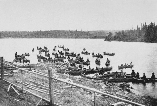 Des familles anicinabek arrivent à Kitcisakik en canot. On y voit une vingtaine de canots dans lesquels sont assises des personnes. On voit la forêt en arrière-plan. Photo en noir et blanc.
