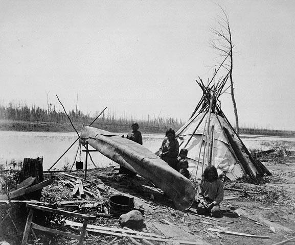 Campement d'été, fabrication de canot. Le site de fabrication de canot est près d'une tente sur le bord du lac. On y voit trois adultes et deux enfants. Photo en noir et blanc.