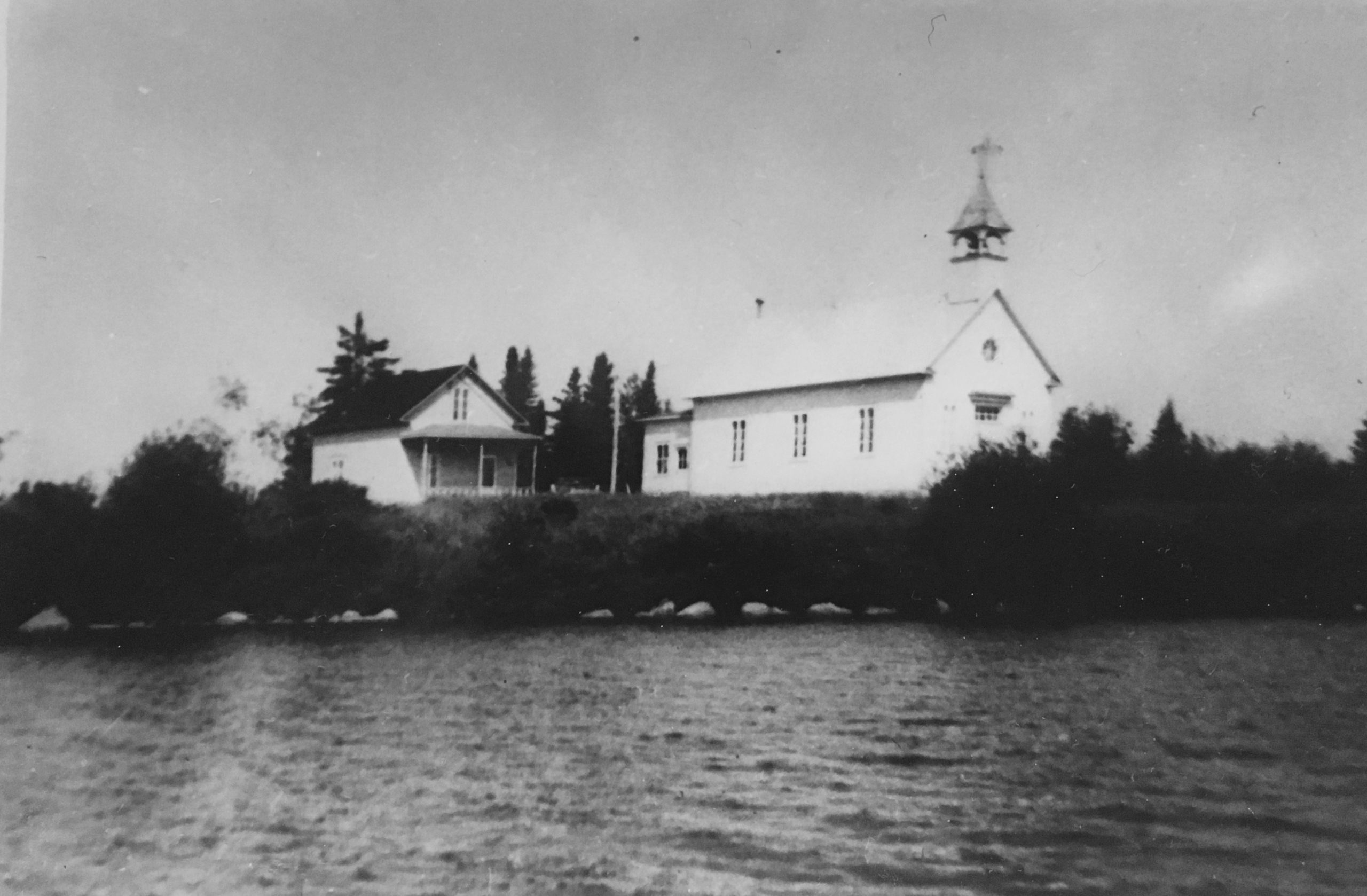 Église de Lac Simon alors qu'elle est située près du Lac Simon. On y voit un autre bâtisse à ses côtés et le lac auquel elle fait face. Photo en noir et blanc.