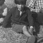 Young Anicinabe boy sitting in the grass near Lake Simon. He is wearing a jacket, shorts, short hair and shoes. A man is on his right and a young man on his left. Black and white picture.