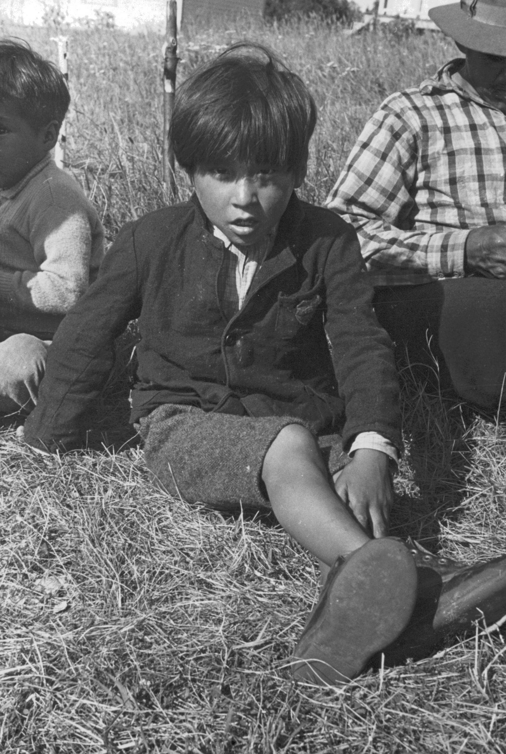 Jeune garçon anicinabe assis dans l'herbe près du Lac Simon. Il porte un veston, des culottes courtes, des cheveux courts et des souliers. Un homme est à sa droite et un jeune homme à sa gauche. Photo en noir et blanc.