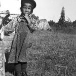 Jeune fille anicinabe près du Lac Simon. Elle porte une robe à manche longue et un béret. Elle porte sa main à sa bouche tout en regardant la caméra. Derrière elle, on y voit une portion de l'église et une grosse roche.  Photo en noir et blanc.