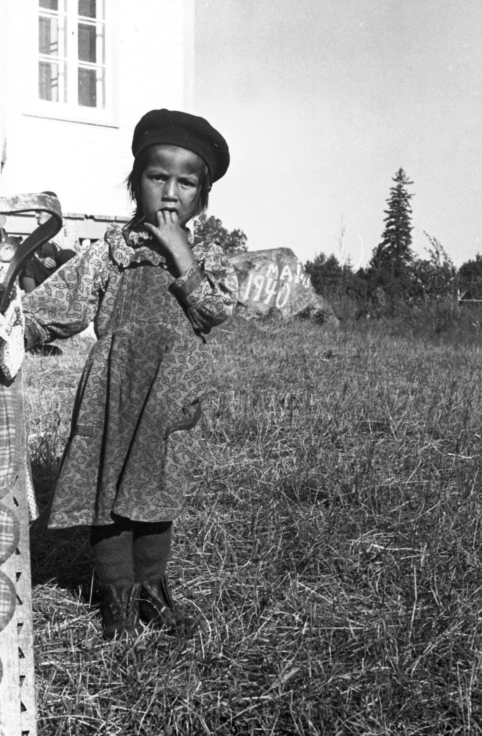 Jeune fille anicinabe près du Lac Simon. Elle porte une robe à manche longue et un béret. Elle porte sa main à sa bouche tout en regardant la caméra. Derrière elle, on y voit une portion de l'église et une grosse roche.  Photo en noir et blanc.