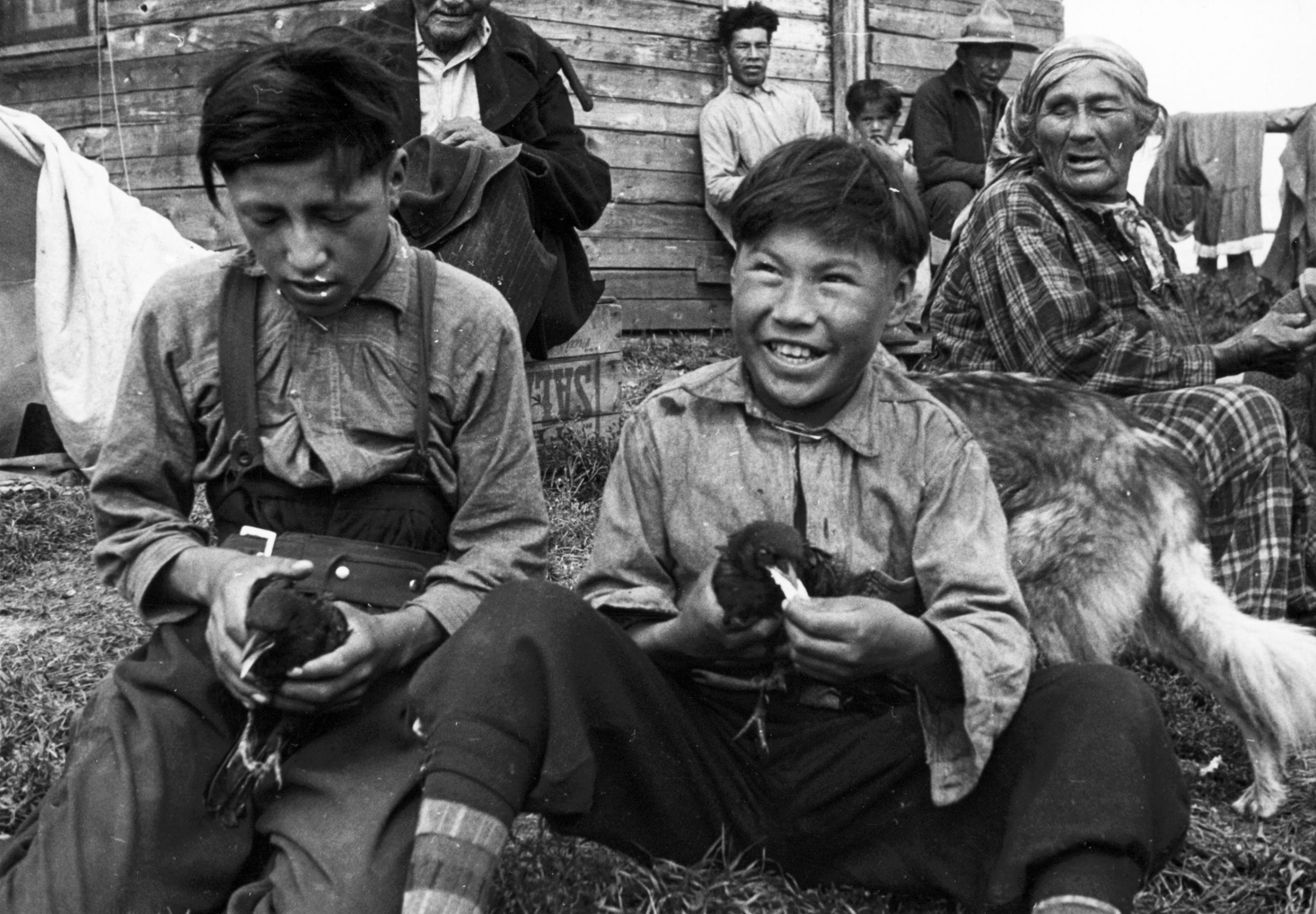 Jeunes anicinabek jouant avec des corneilles qu'ils tiennent dans leurs mains. Ils portent des chemises et des pantalons. Quelques personnes et un chien les entourent. Un bâtiment en planche se dresse au fond. Des tissus sèchent au vent.  Photo en noir et blanc.