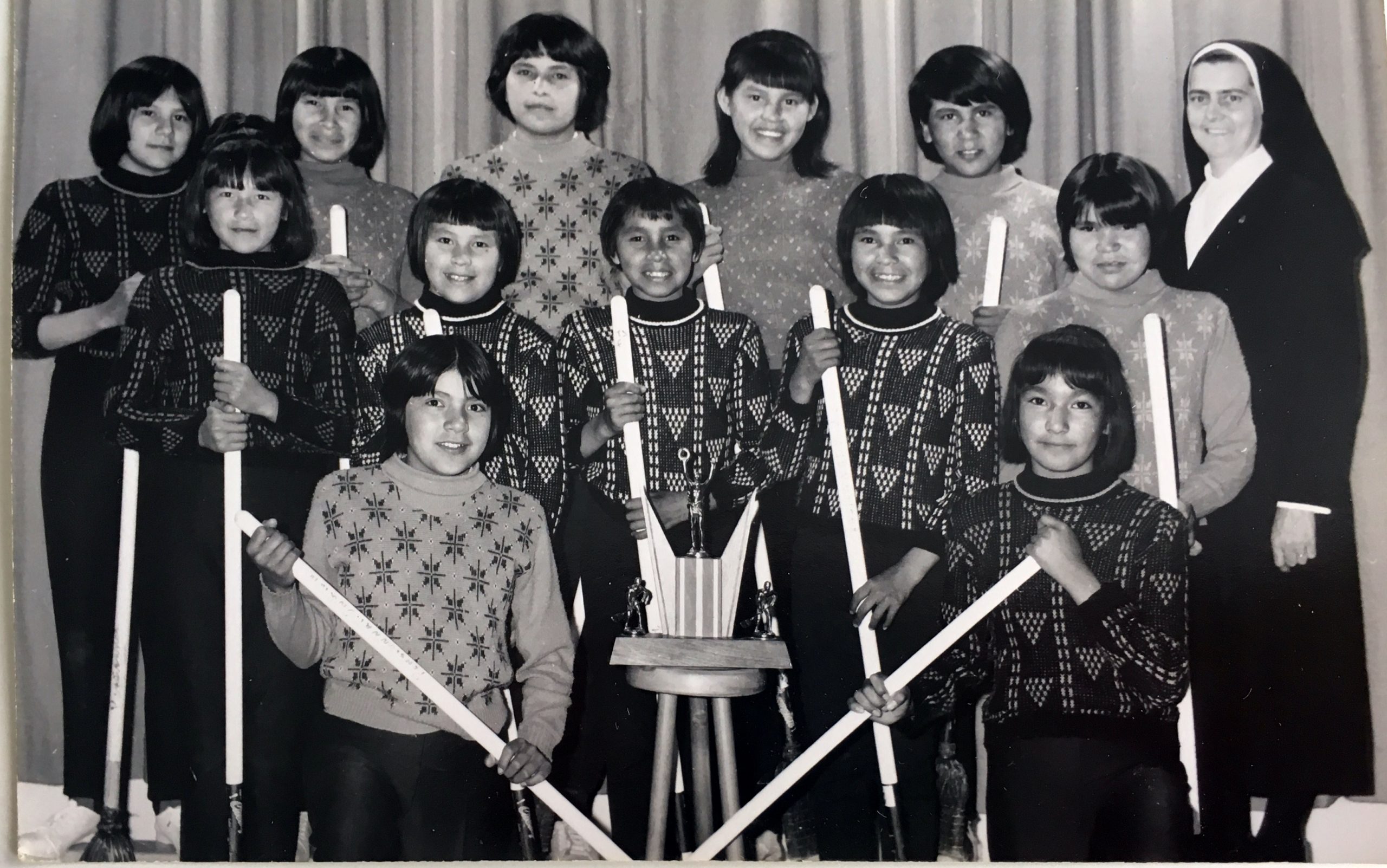 Une équipe féminine de ballon balais au pensionnat d'Amos. Chacune d'elle tient son bâton dans ses mains. Un trophée est au milieu du groupe et une soeur se trouve sur la droite.