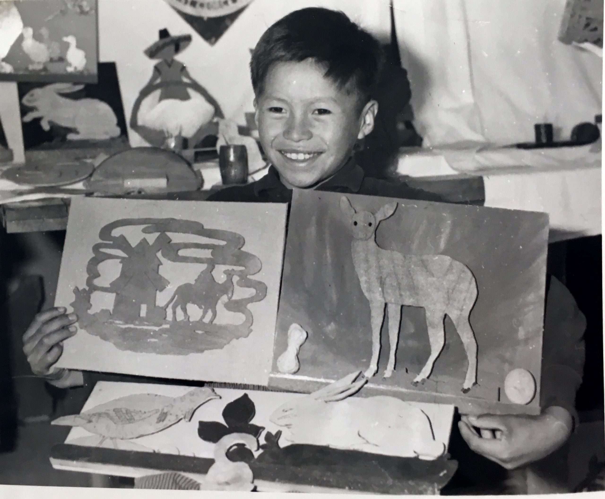 Jeune anicinabe présentant ses oeuvres d'arts au pensionnat d'Amos. Tout sourire, il montre ses gravures qui représentent des animaux notamment. Photo en noir et blanc.