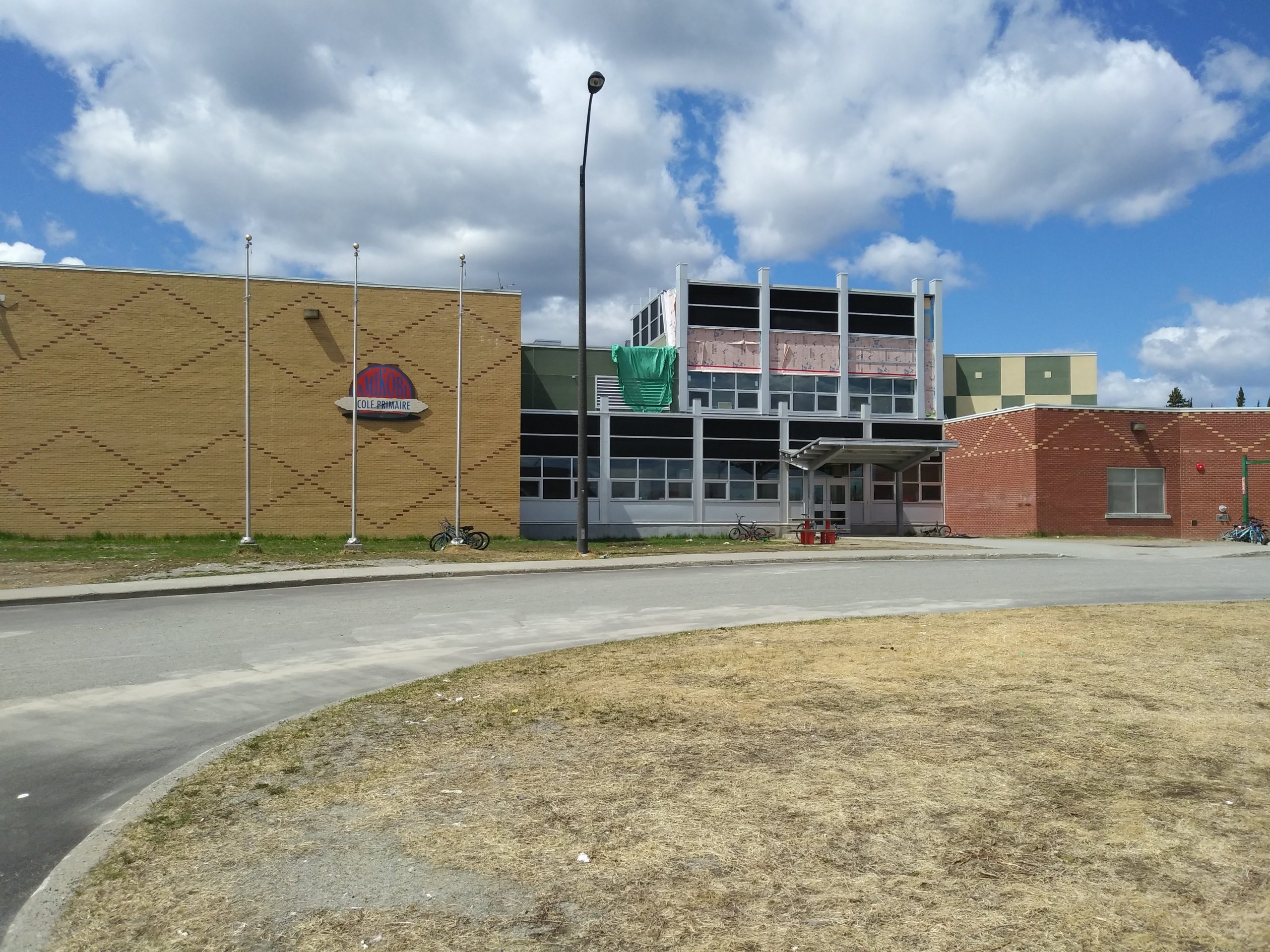 Devanture de l'école primaire de Lac Simon. Une portion est en briques jaunes, l'autre en briques rouges ainsi qu'une portion fenêtrée au centre. On y voit une portion de la route qui mène à l'entrée.  Photo en couleur.