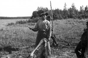 Un homme anicinabe tient un grattoir traditionnel à la main près d'une peau qu'il est en train de travailler. Un autre homme est derrière lui et un à ses côtés. Ils sont près d'un lac. Photo en noir et blanc.