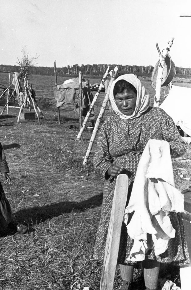 Une femme anicinabe avec une peau tannée dans ses mains. Elle porte une robe longue et un foulard sur sa tête. On voit une portion de campement derrière elle avec des tentes et supports en bois. Photo en noir et blanc.