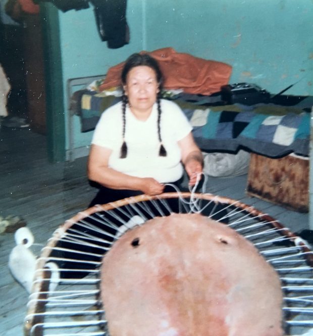 Une femme anicinabe assise au sol est en train de tendre une peau de castor sur un support de bois en forme de cercle à l'aide de corde. Derrière elle, on voit un lit. Photo en couleur.