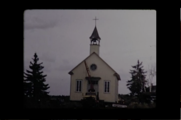 L'église est en train d'être déplacée sur le nouveau site de la communauté. Elle est déplacée par camion sur l'image. On y voit le devant et le clocher. Photo en couleur.