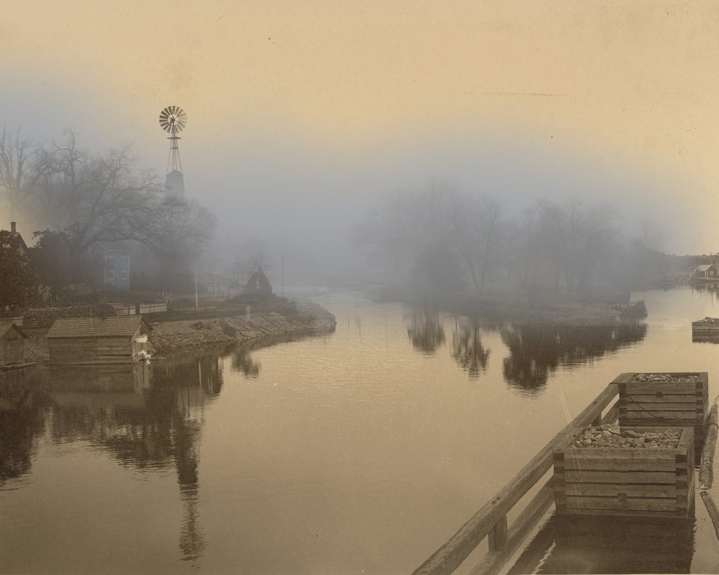  Une photo en noir et blanc d'un canal; dans l'image contemporaine une maison cochère bleue est visible à travers le brouillard.