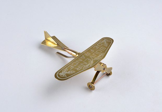 Petit avion en laiton lustré fait avec une cartouche de carabine. Il possède une hélice à l’avant et l’aile porte l’inscription « Souvenir de France. »