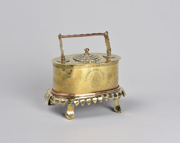 Une petite boîte à bijoux en laiton de forme ovale avec une poignée et quatre pieds. Au sommet se trouve une petite serrure à combinaison richement décorée.