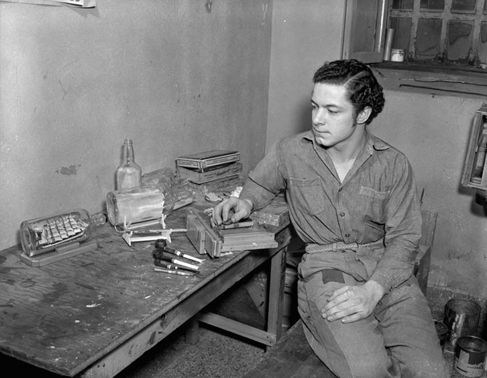 Prisonnier de guerre allemand, assis devant un établi et regardant une de ses créations, un voilier dans une bouteille.