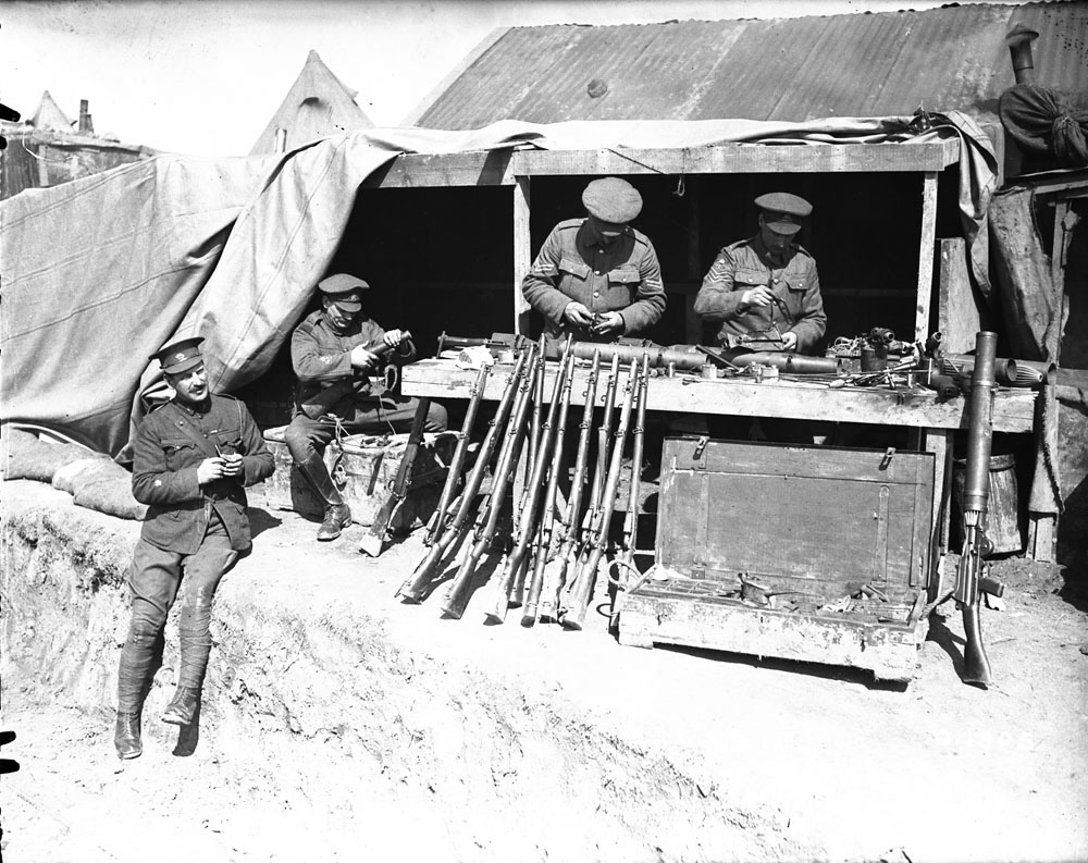Photo en noir et blanc d’une armurerie improvisée. Deux soldats derrière une table étudient divers armements, et deux autres soldats sont présents.