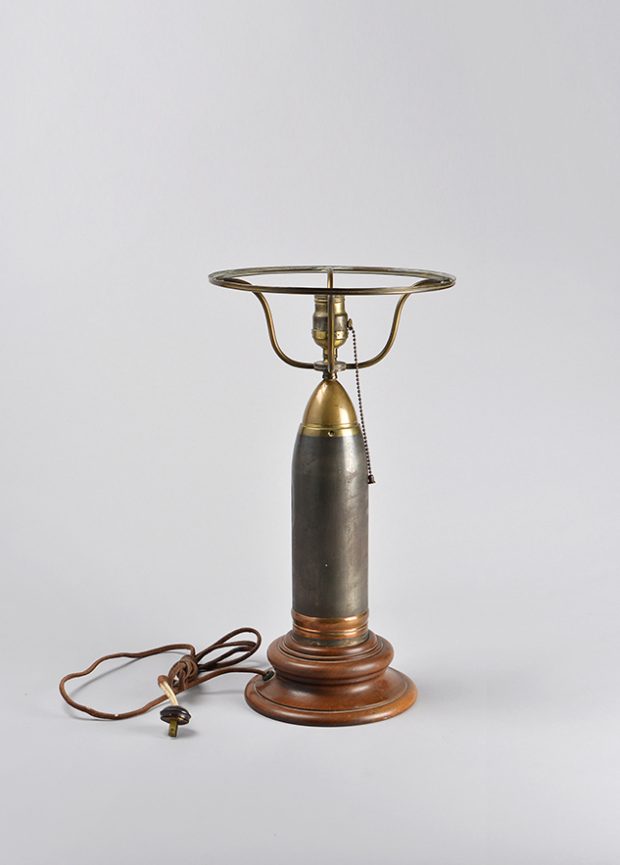 Lampe de table avec base circulaire en bois orné de moulures; le corps est fait d’une douille d’obus au-dessus de laquelle se trouve  une douille électrique entourée d’une armature pour soutenir un abat-jour.