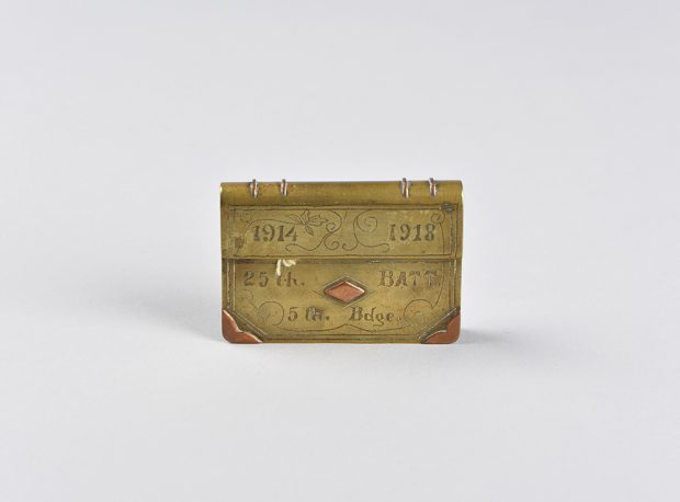 Briquet en laiton et cuivre, en forme de porte-documents miniature. Des dates et d’autres caractères sont gravés, et le couvercle est refermé.