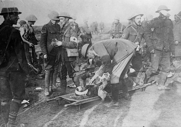 Photo en noir et blanc d’une scène de guerre chaotique. Au centre, sur une civière, un soldat blessé tient fermement un objet-souvenir.