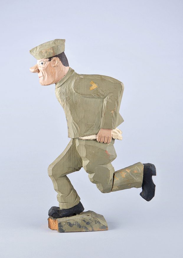 Sculpture sur bois, de style rustique, d’un soldat en pleine course. Il a un uniforme vert, des bottes noires et sa chemise n’est pas rentrée dans son pantalon.