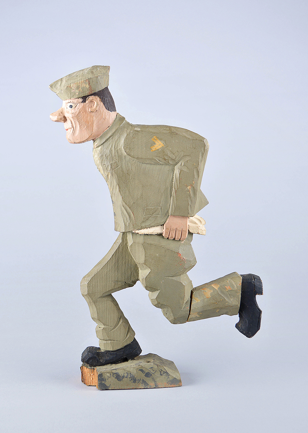 Sculpture sur bois, de style rustique, d’un soldat en pleine course. Il a un uniforme vert, des bottes noires et sa chemise n’est pas rentrée dans son pantalon.