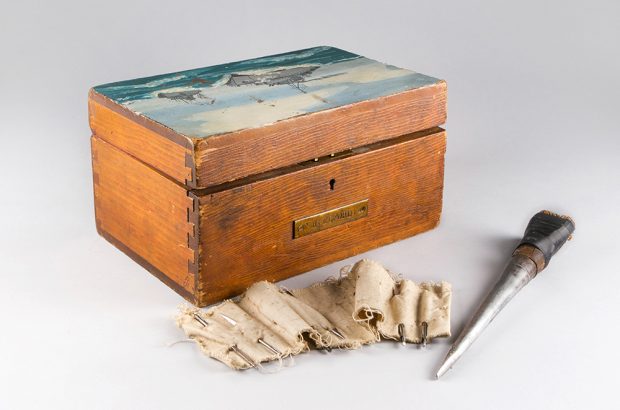 Boîte de bois, appelée un « nécessaire de marin, » avec deux vaisseaux de guerre  peints sur le couvercle. Devant la boîte se trouve une série d’aiguilles enfilées sur un bout de toile très usée, ainsi qu’un épissoir improvisé.