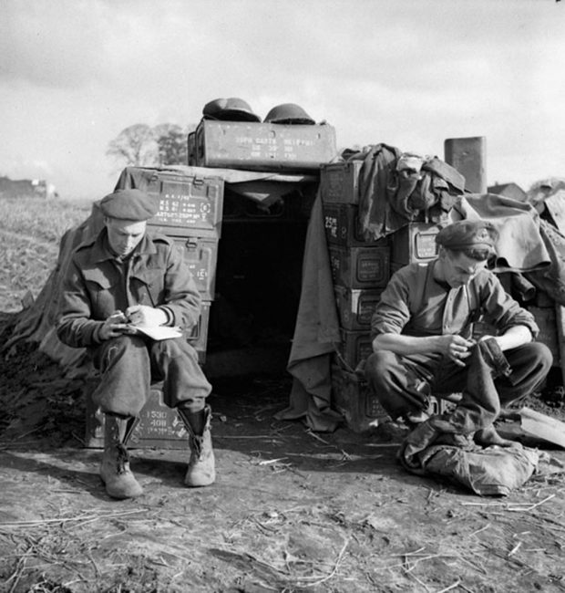 Photo en noir et blanc de deux bombardiers devant un abri improvisé, en lisière d’un champ. L’un écrit et l’autre se livre à un travail d’artisanat.