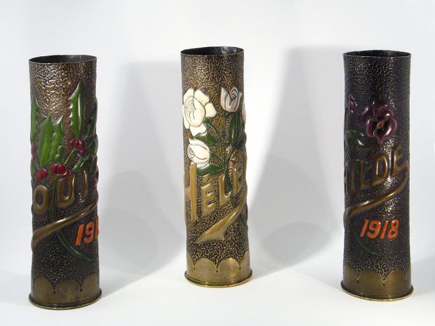 Trois douilles d’obus, transformées en vases, avec des motifs de plantes, des noms et des dates estampés sur la surface. Les plantes et les noms sont en couleur.