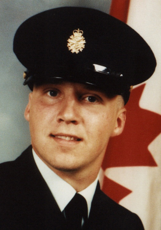 Gros plan en couleurs d’un soldat souriant et en uniforme, devant un drapeau canadien.