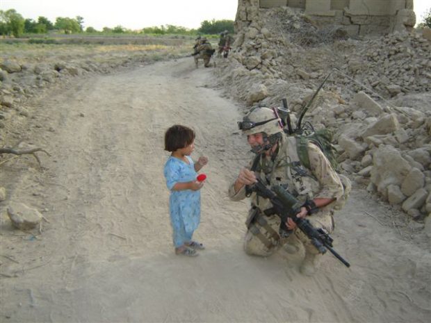 Photo en couleurs d’un soldat à genoux, en tenue de combat, donnant une poupée Izzy à un très jeune enfant.