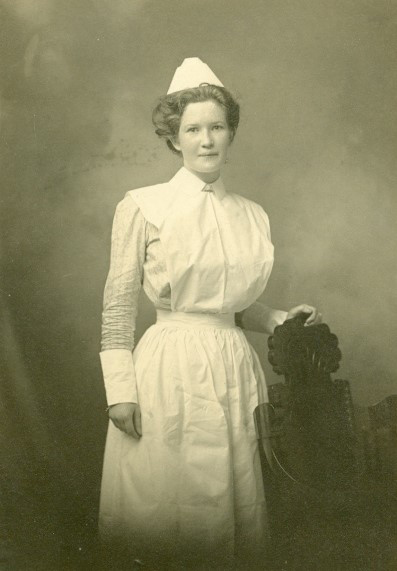 Photo sépia d’une infirmière en uniforme, la main posée sur le dos d’une chaise abondamment sculptée.