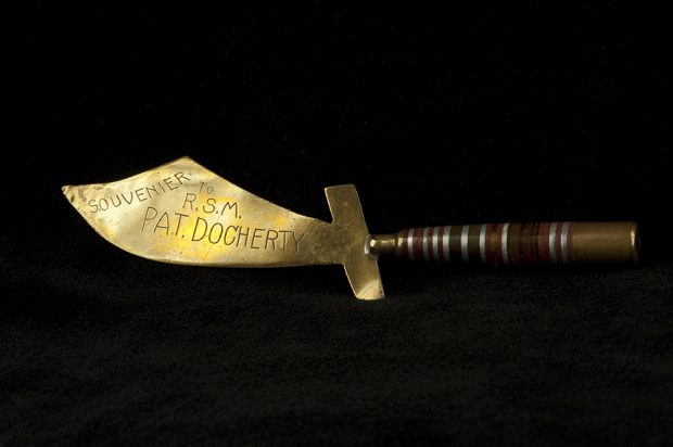 Photo en couleurs d’un ouvre-lettres en forme de sabre. La poignée est faite d’une cartouche, attachée à une lame de laiton lustré, portant une inscription gravée.