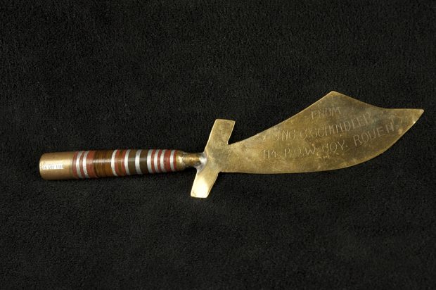 Photo en couleurs d’un ouvre-lettres en forme de sabre. La poignée est faite d’une cartouche, attachée à une lame de laiton lustré, portant une inscription gravée.
